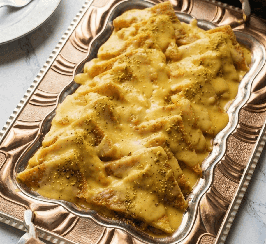 Shahi Tukra, featuring golden-brown fried bread slices layered on a long, ornate copper-toned serving tray with intricate handles. The bread is generously smothered in a thick, creamy saffron-infused milk sauce and finished with a delicate dusting of crushed green pistachios.